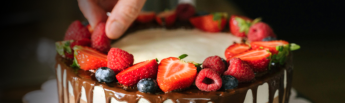 A hand placing a raspberry atop a multi-layered chocolate cake adorned with a variety of berries and a chocolate fudge drizzle